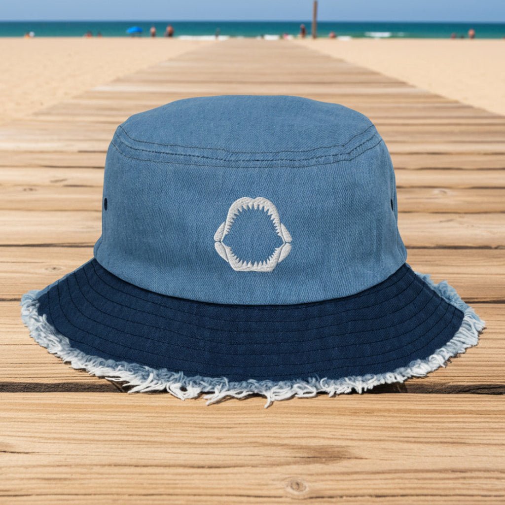 A two-tone blue bucket hat made of washed light blue denim on the crown and a darker navy blue denim brim with frayed edges. A white embroidered shark-tooth motif is stitched on the front center of the crown.