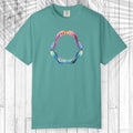 A green short-sleeve t-shirt with a colorful graphic of a shark mouth on the chest, displayed flat against a white wood plank background.