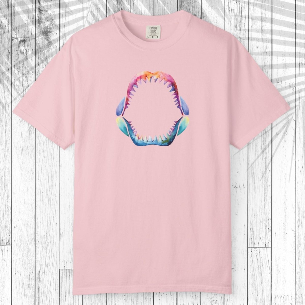 A light pink short-sleeve crewneck t-shirt featuring a colorful graphic of an open shark mouth with sharp teeth on the chest, displayed flat against a white wood plank background.