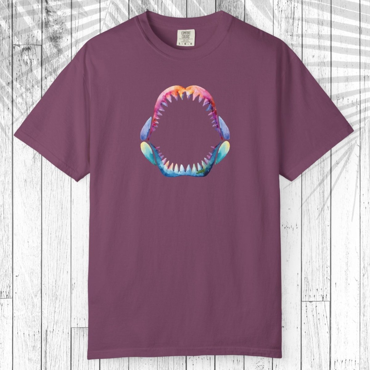 A short-sleeve crewneck t-shirt in a dusty plum purple color, featuring a colorful graphic of a shark's open mouth with sharp teeth on the chest. The shirt is displayed flat against a white wood plank background.
