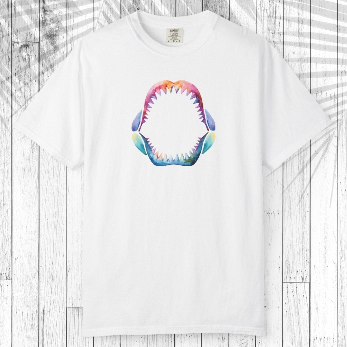 A white short-sleeve crewneck t-shirt featuring a colorful graphic of an open shark mouth with sharp teeth, displayed flat against a white wood plank background with a palm leaf overlay.