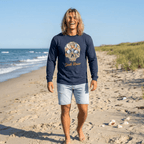 A man walking on a sandy beach wearing a navy blue long-sleeve t-shirt with a colorful skull graphic and the words 'Shell Rising' printed on the front, paired with light denim shorts.