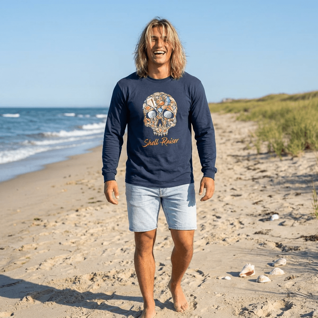 A man walking on a sandy beach wearing a navy blue long-sleeve t-shirt with a colorful skull graphic and the words 'Shell Rising' printed on the front, paired with light denim shorts.