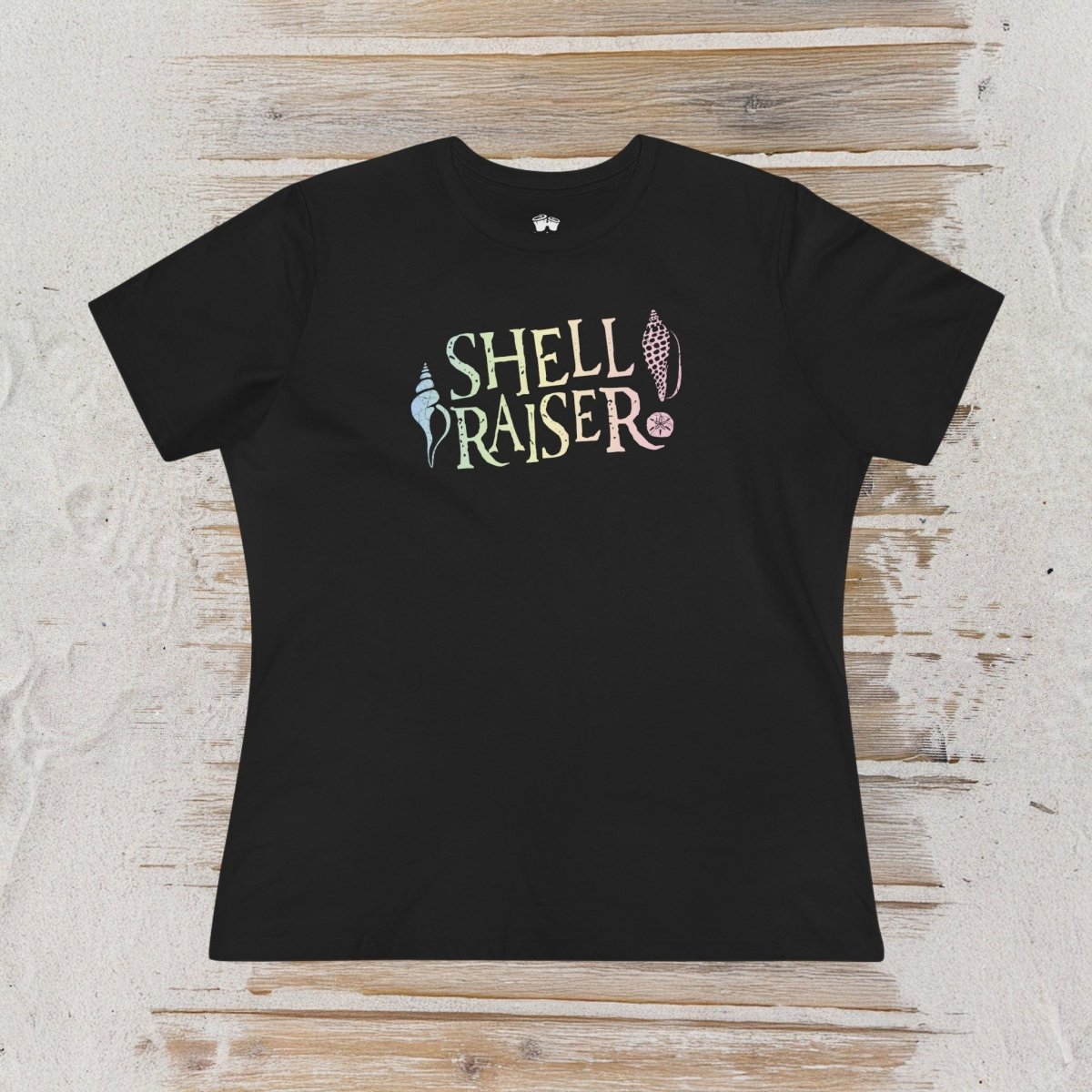 A navy blue short-sleeve t-shirt with a colorful graphic reading 'SHELL RAISER' in stylized lettering, laid flat on a light wood plank surface.