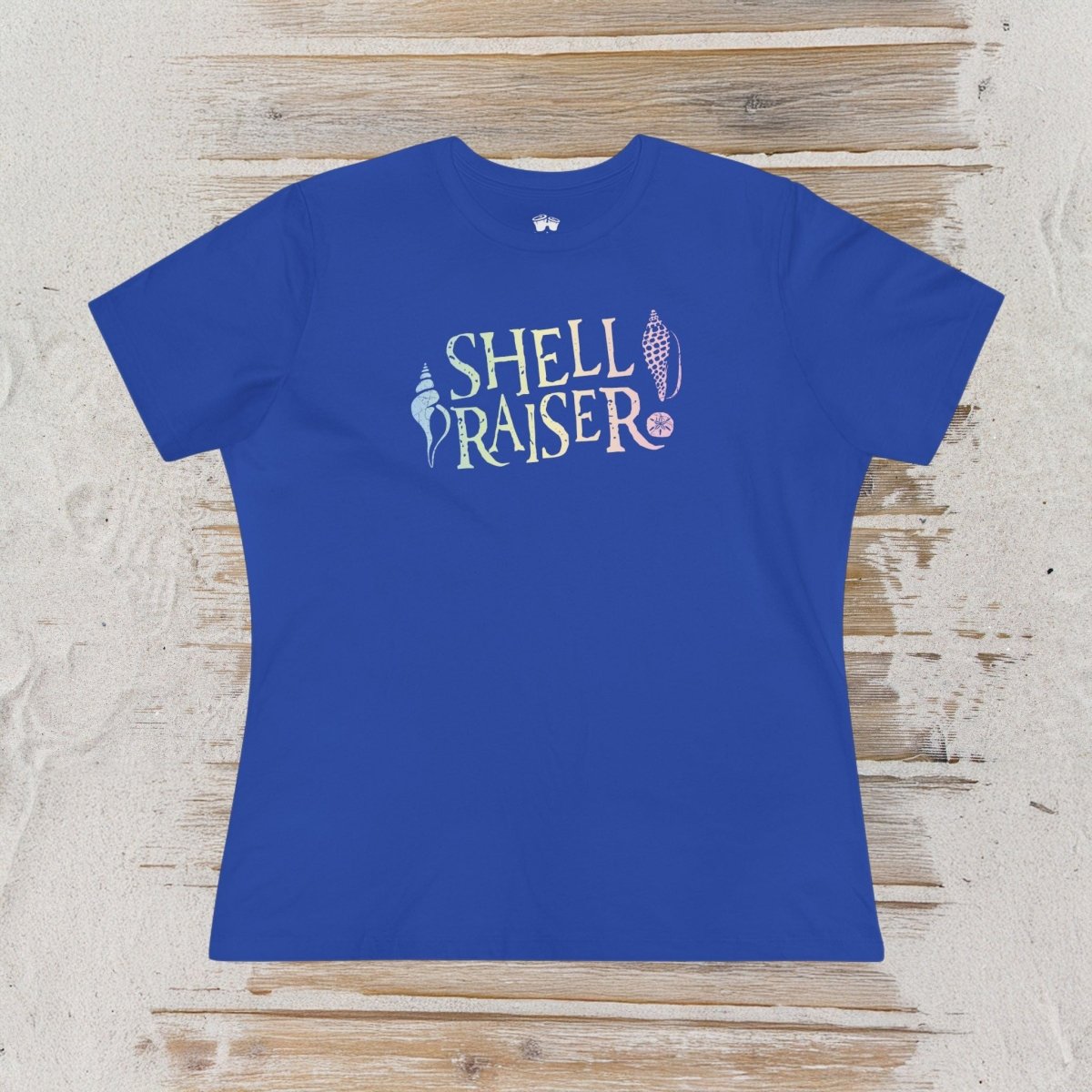 A royal blue short-sleeve t-shirt with a graphic reading 'SHELL RAISER' in a stylized multicolor font, laid flat on a light-colored wooden plank surface.