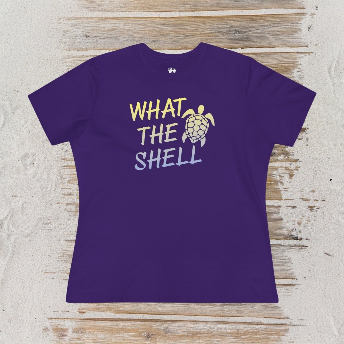 A purple short-sleeve t-shirt with a graphic reading 'WHAT THE SHELL' in yellow and light blue text next to a sea turtle illustration, laid flat on a light wood plank background.