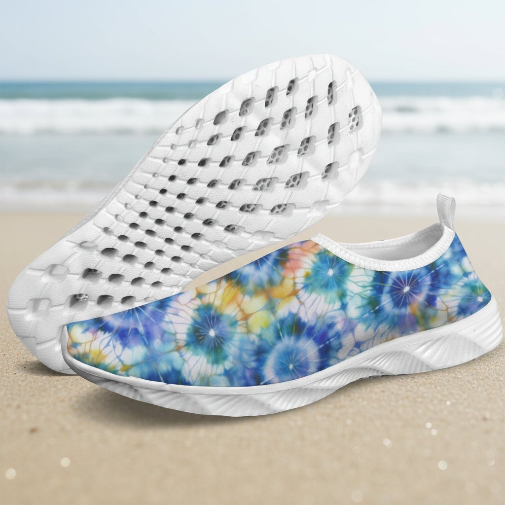 A pair of slip-on water shoes with a blue, yellow, and white tie-dye pattern on the upper, shown faded beach background. One shoe is positioned to show the side pattern, while the other is angled to display the textured white sole.
