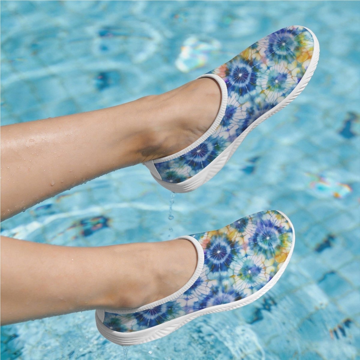 A pair of slip-on water shoes with a blue, purple, and yellow tie-dye floral pattern, shown being worn on feet floating above a swimming pool.