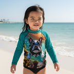 A young girl stands on Siesta Key beach wearing a teal long-sleeve swim rash guard with a large graphic print of a French bulldog wearing a scuba mask. The background shows the blue ocean and sky.