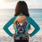A tween girl standing on a beach with her back to the camera, wearing a teal long-sleeve rash guard with a large graphic print of a French Bulldog wearing scuba diving goggles on the back.