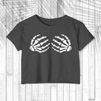 A dark charcoal gray short-sleeve crop top t-shirt featuring a white graphic of two skeletal hands on the chest, displayed flat against a white wood plank background.
