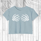 A light blue short-sleeve crop top t-shirt featuring a white graphic of two skeletal hands on the chest, laid flat on a white wood plank background.