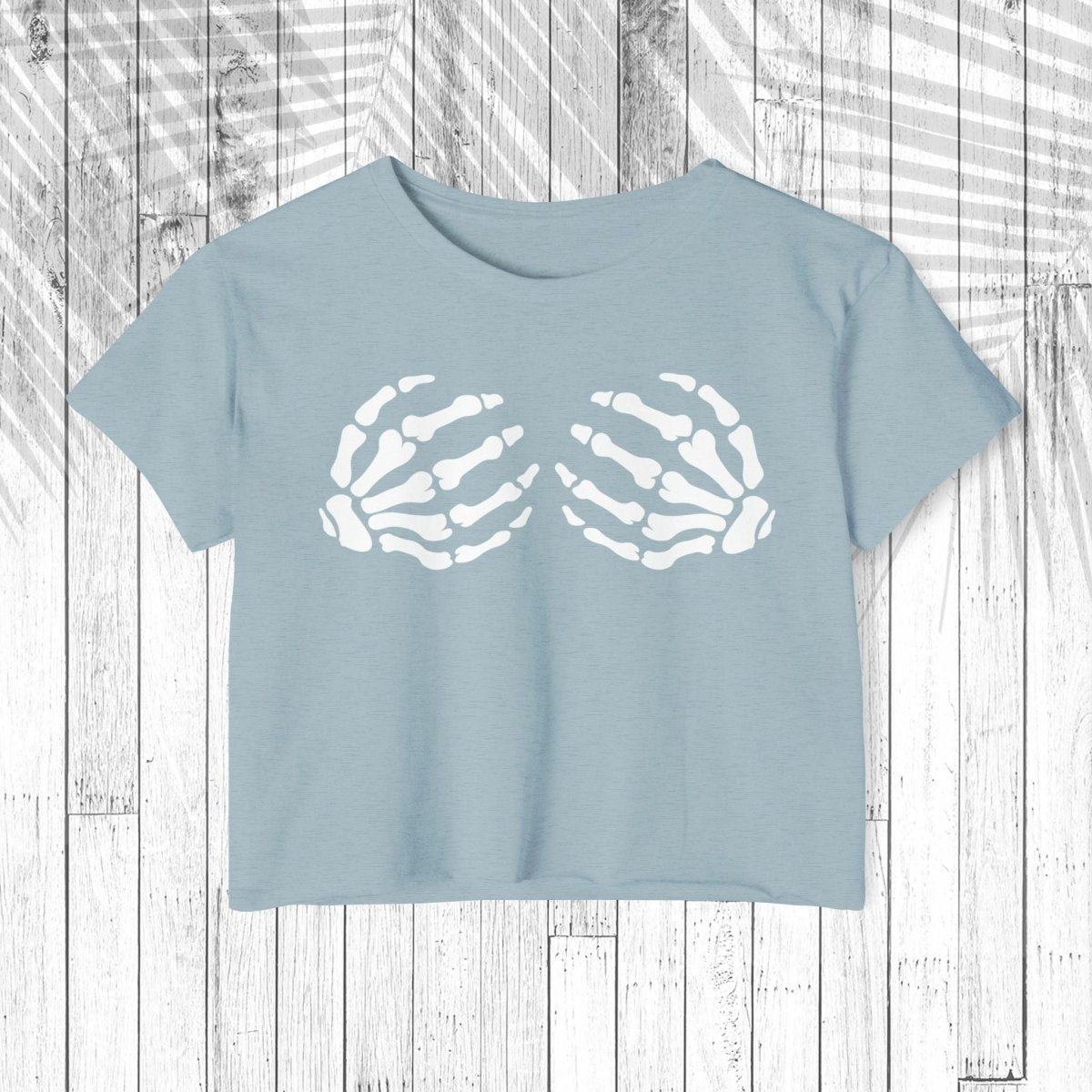 A light blue short-sleeve crop top t-shirt featuring a white graphic of two skeletal hands on the chest, laid flat on a white wood plank background.