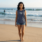 A woman stands on a sandy beach wearing a heathered dark gray sleeveless, Hang Loose Shaka Sign tank top with a white shaka hand made with skeleton fingers on the chest, paired with denim shorts. The ocean waves and surfers are visible in the background.