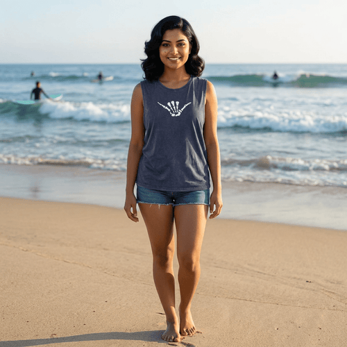 A woman stands on a sandy beach wearing a heathered dark gray sleeveless, Hang Loose Shaka Sign tank top with a white shaka hand made with skeleton fingers on the chest, paired with denim shorts. The ocean waves and surfers are visible in the background.