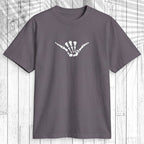 A dark gray crewneck t-shirt featuring a light teal graphic of a dog standing on a surfboard across the chest, displayed flat against a white wood plank background with a palm leaf shadow overlay.