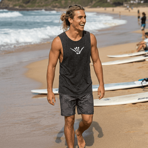 A man walking on a sandy beach wearing a dark charcoal gray sleeveless tank top with a white skeleton hand logo and matching dark gray swim trunks. Surfboards are lined up on the beach behind him, and the ocean waves are visible in the background.