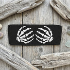 A black rectangular headband featuring a white graphic print of two skeletal hands facing each other, shown against a rustic wood background.