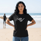 A woman standing on a sandy beach wearing a black short-sleeve t-shirt with a white skeleton hand shaka graphic on the chest, paired with blue jeans. The background shows the ocean and a blurred surfer.