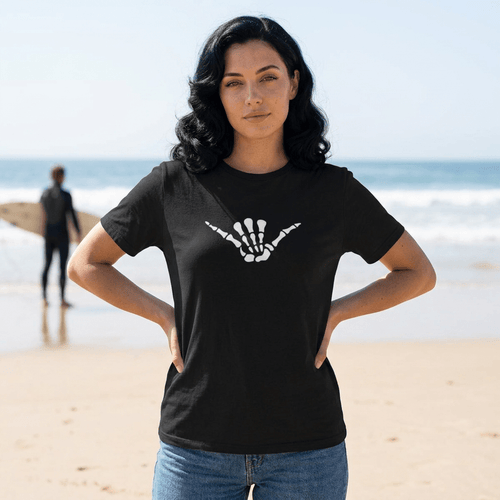 A woman standing on a sandy beach wearing a black short-sleeve t-shirt with a white skeleton hand shaka graphic on the chest, paired with blue jeans. The background shows the ocean and a blurred surfer.