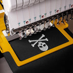 Close-up of an embroidery machine stitching a silver skull and crossbones design onto dark navy fabric, with the yellow and white machine body visible.