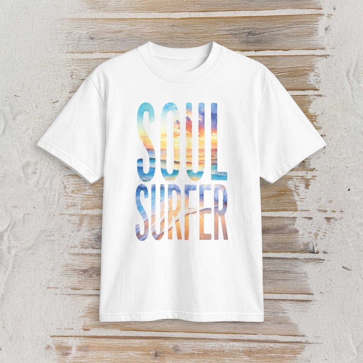 A white short-sleeve crewneck t-shirt with 'SOUL SURFER' printed in a large, colorful watercolor-style font on the front, laid flat on a rustic light wood background.