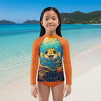 A young girl stands on a sandy beach in  St. Johns wearing a long-sleeve swim shirt with bright orange sleeves and a colorful underwater pufferfish graphic on the front