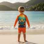 A young child stands on a sandy beach in st john's virgin islands. facing the ocean, wearing a long-sleeve swim shirt with a colorful puffer fish graphic on the back and bright orange swim shorts.