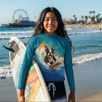 A smiling young tween stands on a Santa Monica Beach, wearing a fitted long-sleeve Youth Rash Guard with a printed graphic of a dog surfing, with a California pier and Ferris wheel visible in the background.