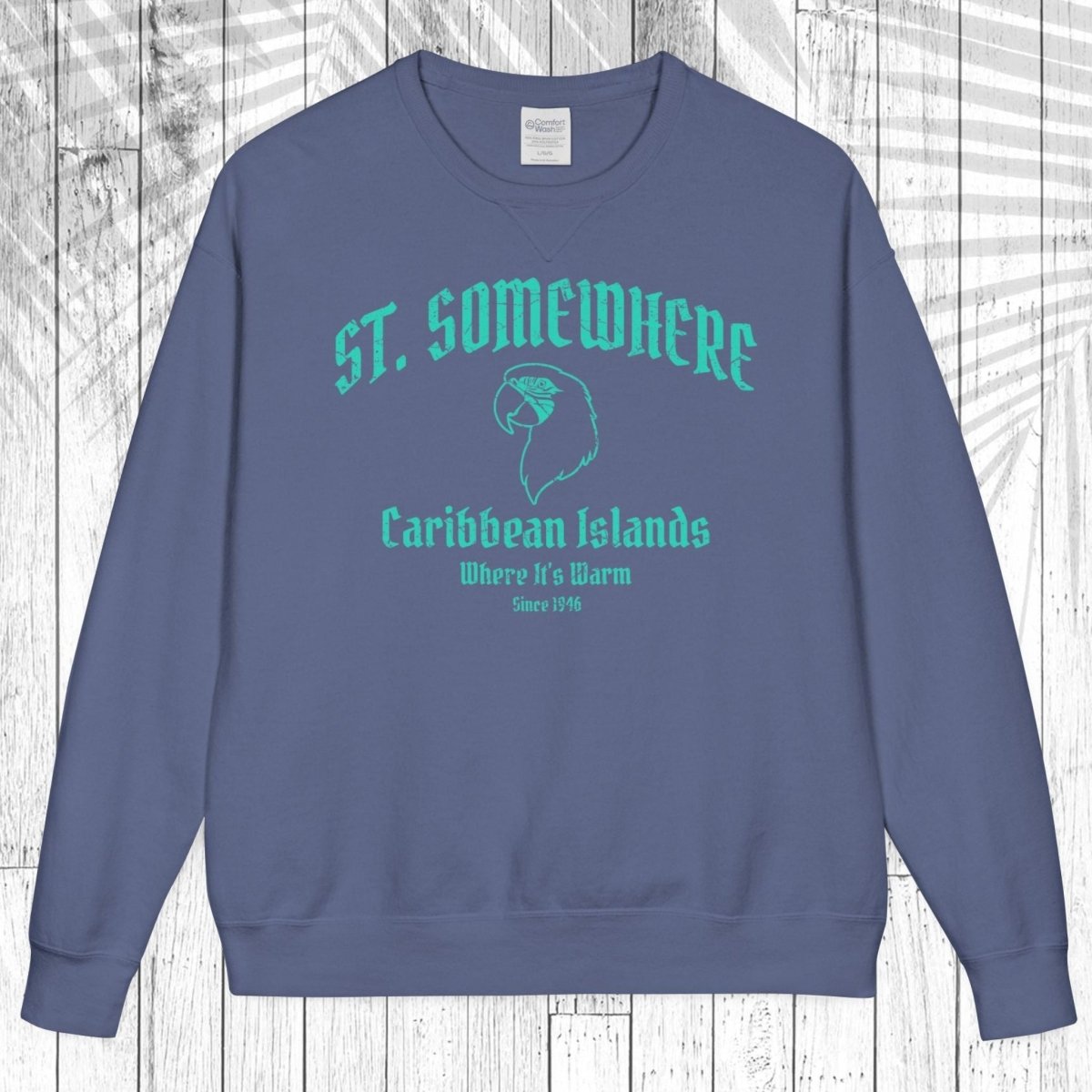 A slate blue crewneck sweatshirt with a teal graphic print reading 'ST. SOMEWHERE Caribbean Islands' and 'Where It's Warm Since 1996' on the front, displayed flat against a white wood plank background.
