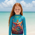 A young girl with long red hair stands on a beach wearing a long-sleeve swim shirt with a colorful cartoon starfish and coral reef graphic on the front.