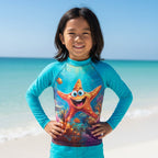 A smiling young girl stands on Starfish Point wearing a bright turquoise long-sleeve rash guard with a colorful underwater print featuring a starfish and coral. The background shows the blue ocean and sky.