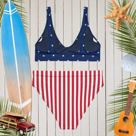 Navy bikini top with white stars and red striped bottom on a wooden background with beach-themed decorations.