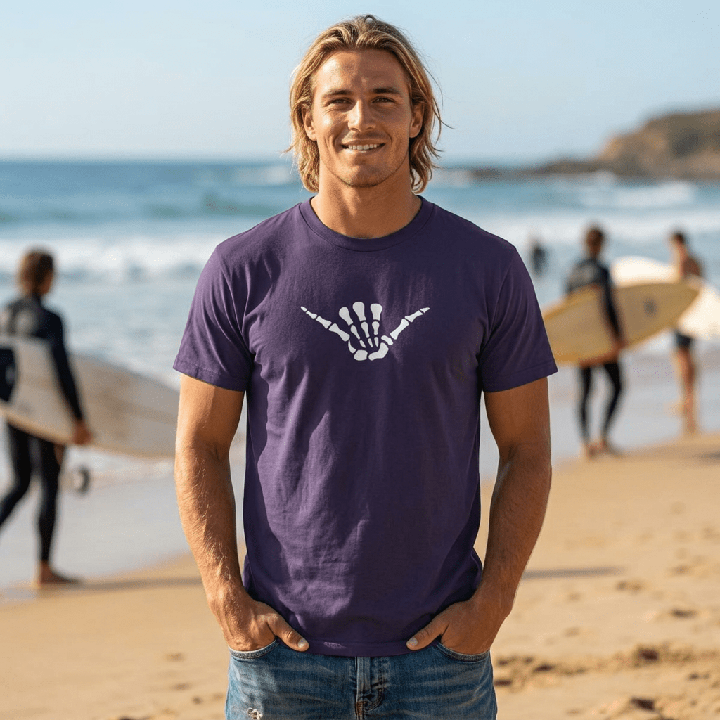 A smiling man with blonde hair stands on a sandy beach wearing a dark purple Skeleton Shaka T-Shirt with a white shaka hand graphic on the chest, with surfers and the ocean blurred in the background.