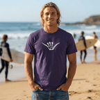 A smiling man with blonde hair stands on a sandy beach wearing a dark purple Skeleton Shaka T-Shirt with a white shaka hand graphic on the chest, with surfers and the ocean blurred in the background.