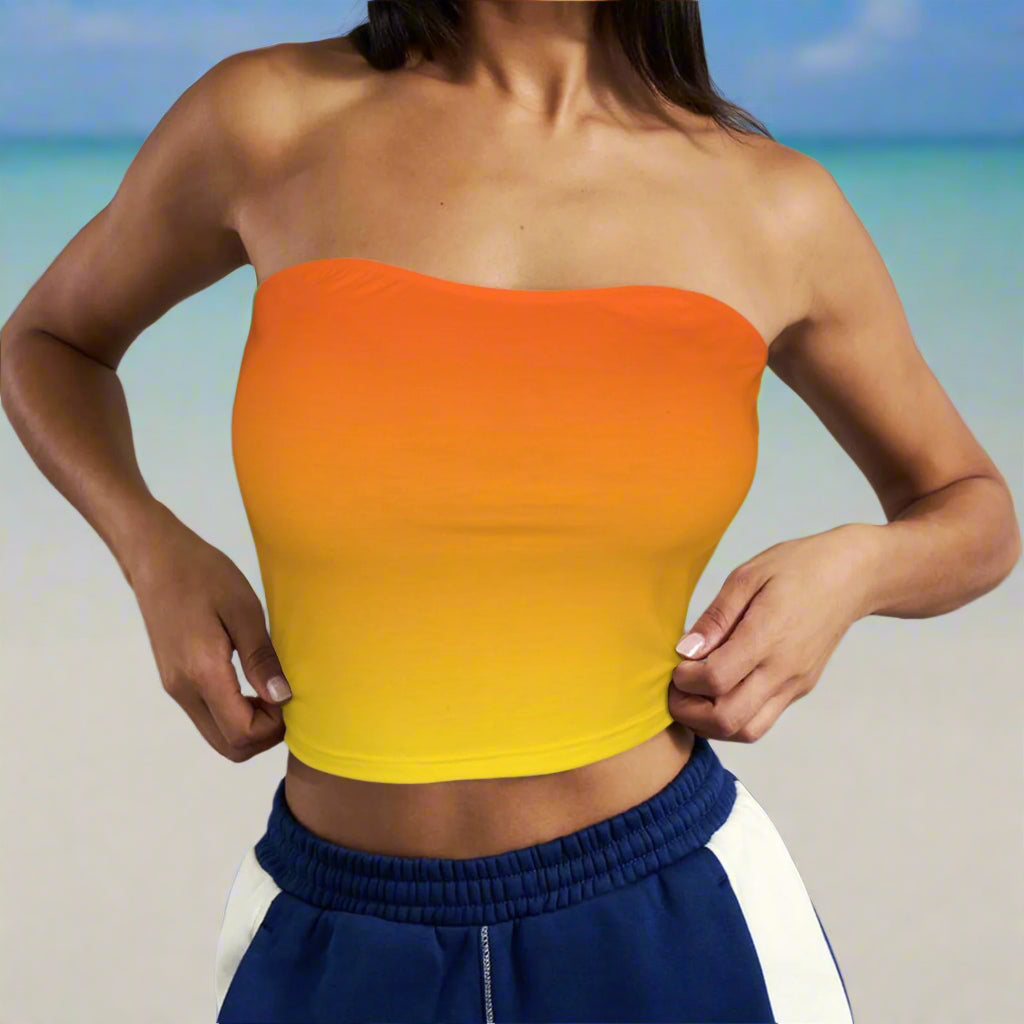 Swimsuit model wearing a gradient orange to yellow strapless top with blue and white pants against a beach backdrop