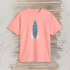 A coral pink short-sleeve t-shirt with a graphic of a blue surfboard containing white handwritten text and white floral accents, displayed flat on a rustic light wood background.