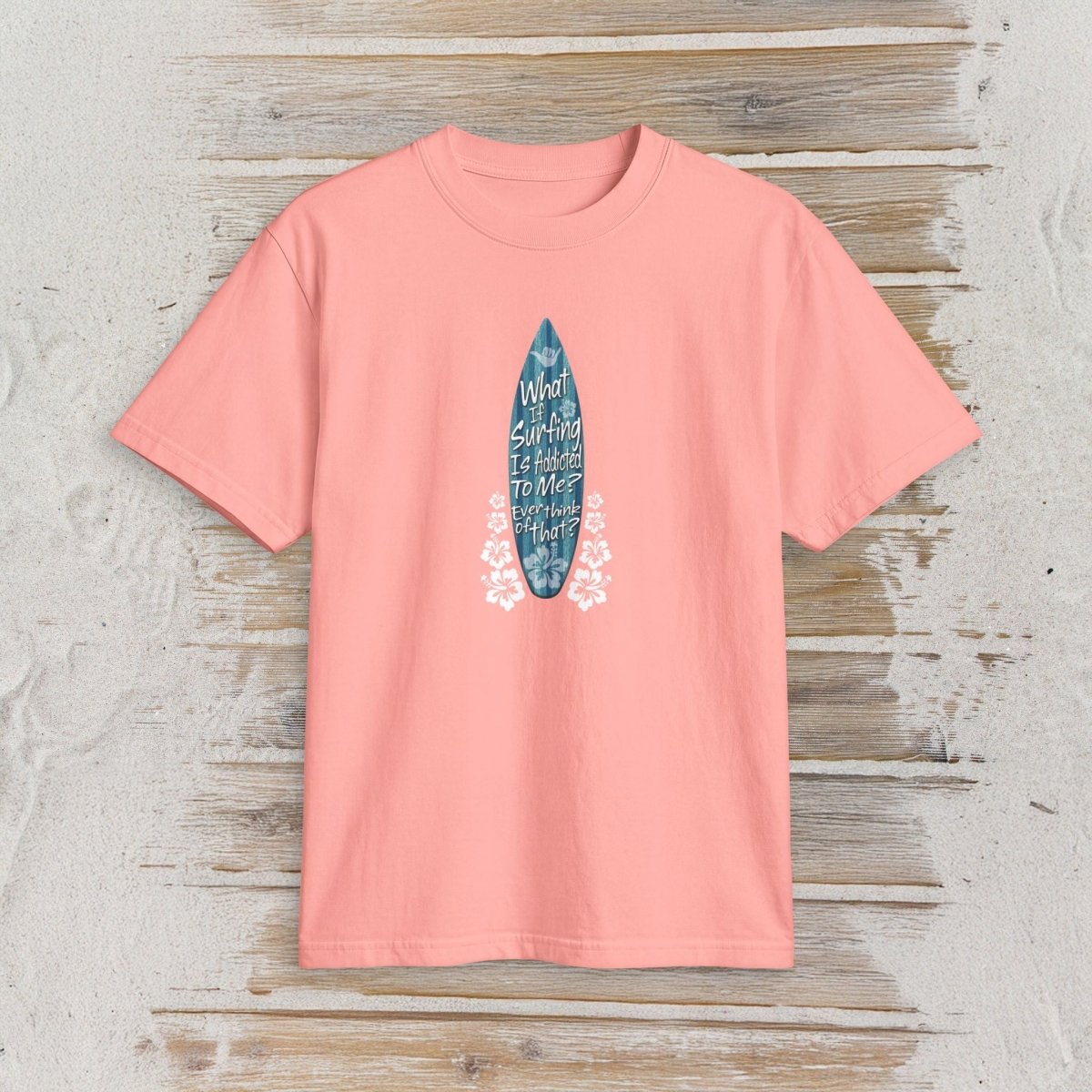 A coral pink short-sleeve t-shirt with a graphic of a blue surfboard containing white handwritten text and white floral accents, displayed flat on a rustic light wood background.