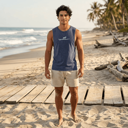 A man standing on a sandy beach wearing a heathered navy sleeveless surfer's tank top with a light gray frenchton bulldog on a surgboard graphic and beige shorts.