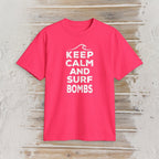A bright pink short-sleeve t-shirt with a white graphic print reading 'KEEP CALM AND SURF BOMBS' and a wave icon, displayed flat on a light wood plank background.