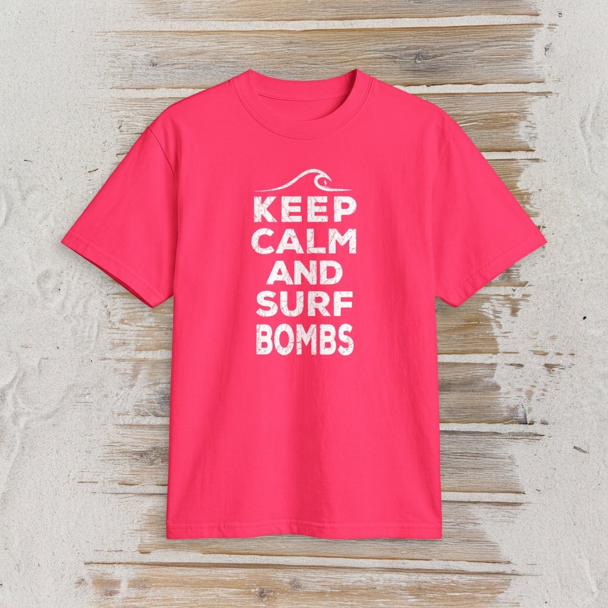 A bright pink short-sleeve t-shirt with a white graphic print reading 'KEEP CALM AND SURF BOMBS' and a wave icon, displayed flat on a light wood plank background.