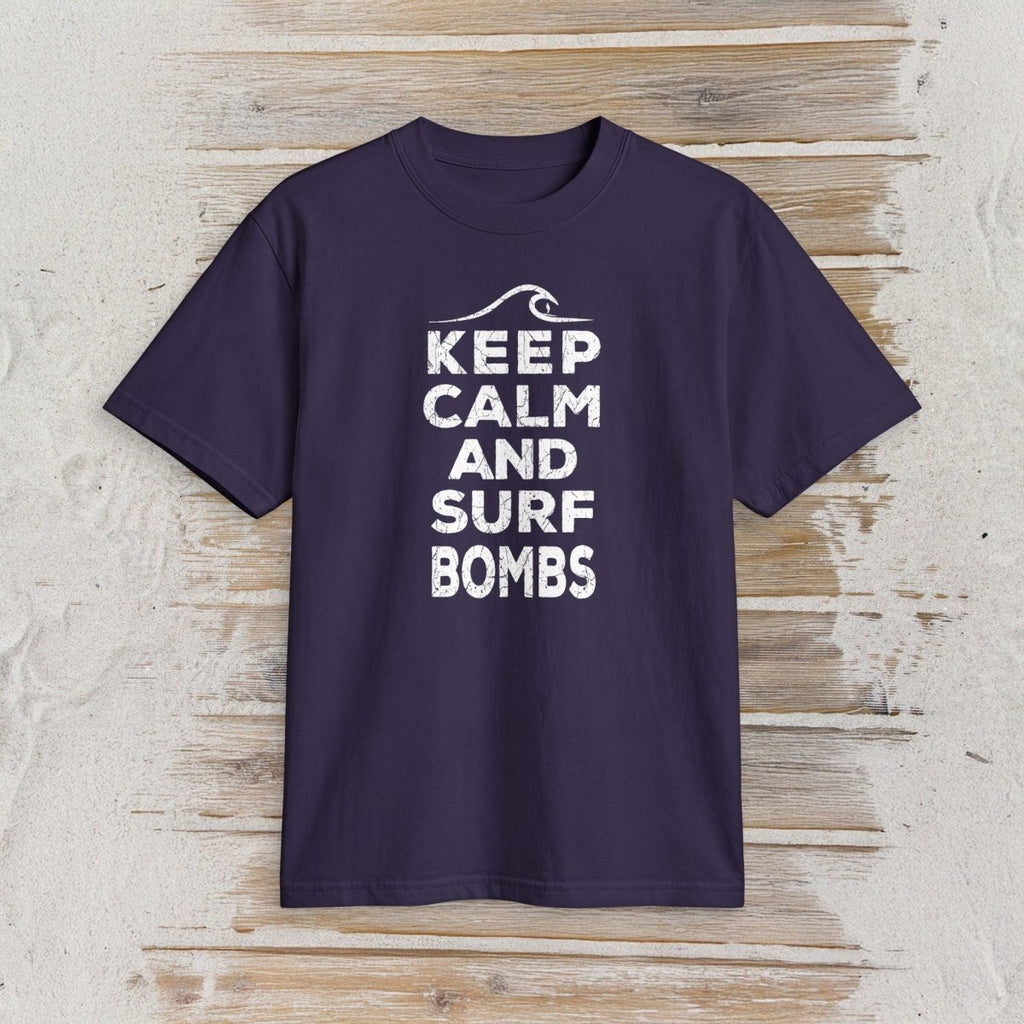 A dark purple short-sleeve t-shirt with a white graphic print reading 'KEEP CALM AND SURF BOMBS' and a wave icon, displayed flat against a light wood plank background.
