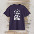 A dark purple short-sleeve t-shirt with a white graphic print reading 'KEEP CALM AND SURF BOMBS' and a wave icon, displayed flat against a light wood plank background.