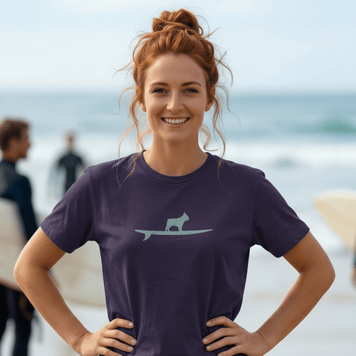 A woman with red hair in a bun stands on a beach wearing a dark purple t-shirt with a light green cat on a surfboard graphic, paired with blue jeans. Surfers are visible in the blurred background.