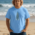 A man with blonde hair stands on a sandy beach wearing a light blue Shaka Wear t-shirt with a graphic of a surfboard and the text 'What Gets You Up Here Won't Get You Down Here'.