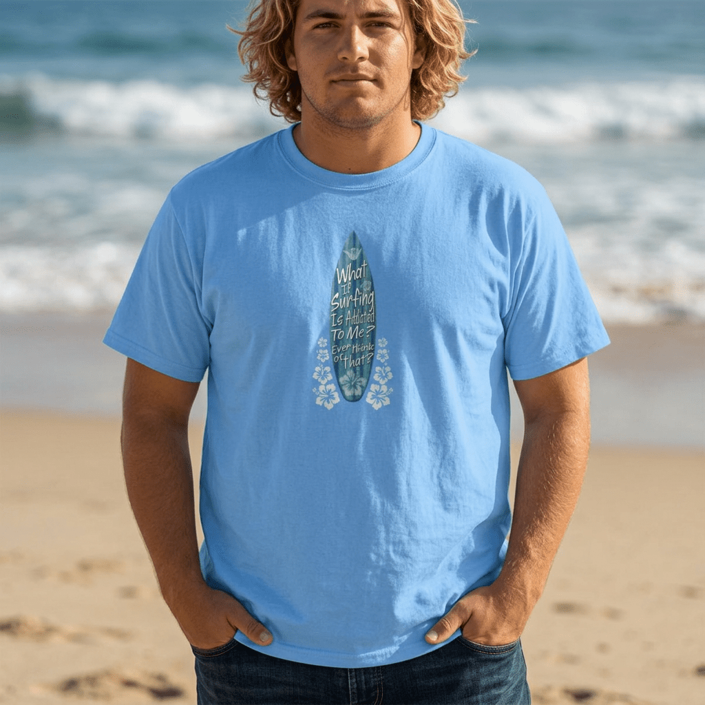 A man with blonde hair stands on a sandy beach wearing a light blue Shaka Wear t-shirt with a graphic of a surfboard and the text 'What Gets You Up Here Won't Get You Down Here'.