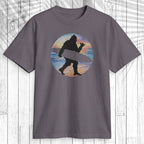 A dark gray crewneck t-shirt featuring a circular graphic of a black silhouette of a Bigfoot-like figure carrying a surfboard, set against a background of blue, orange, and yellow horizontal stripes. The shirt is displayed flat on a white wood panel background.