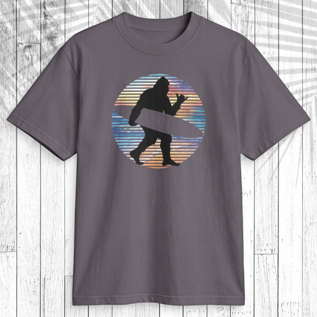 A dark gray crewneck t-shirt featuring a circular graphic of a black silhouette of a Bigfoot-like figure carrying a surfboard, set against a background of blue, orange, and yellow horizontal stripes. The shirt is displayed flat on a white wood panel background.