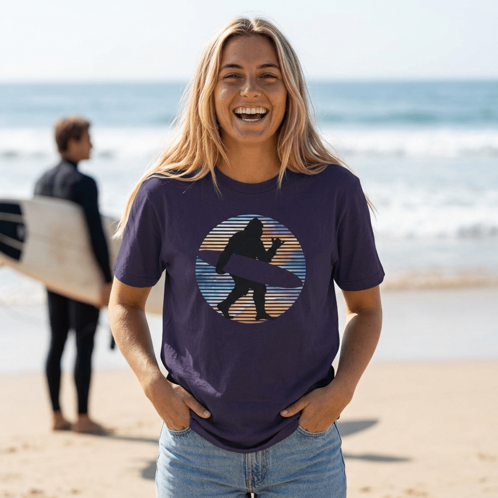 A woman smiling on a beach wearing a dark purple t-shirt with a circular graphic of a silhouetted sasquatch carrying a surfboard flashing a shaka sign against a striped sunset background. A blurred surfer is visible in the background near the ocean.