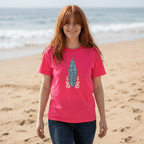 A woman with red hair stands on a sandy beach wearing a bright pink Shaka Wear t-shirt with a blue and pink surfboard graphic, paired with dark blue jeans.